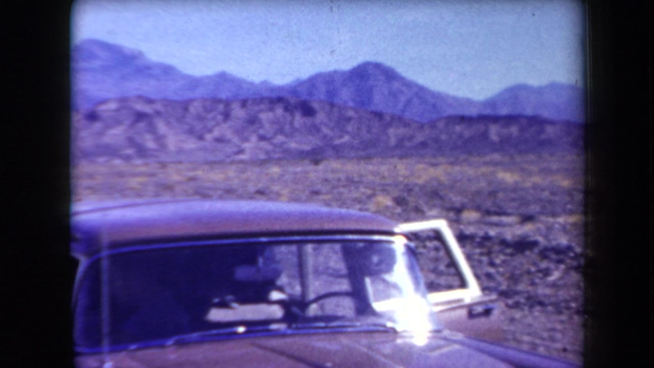 1958 Arizona home-movie frame showing a classic sedan beside a remote desert road with rocky terrain and distant mountains.