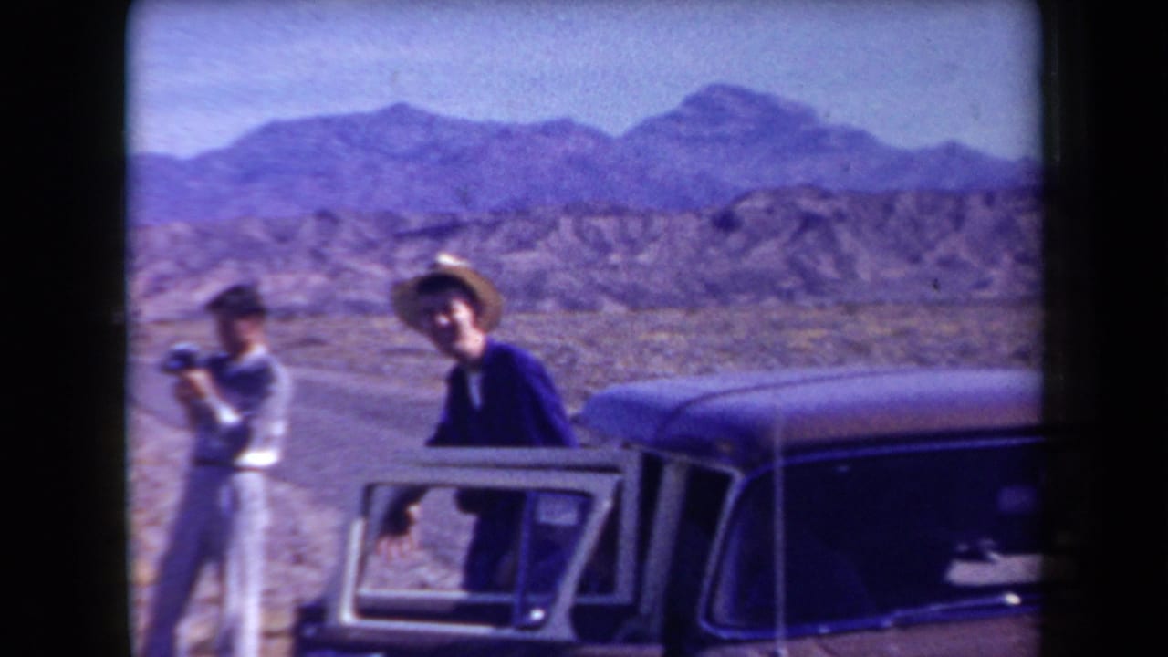 Two adult men pause beside a classic sedan on an Arizona desert road, with one holding a camera and mountains beyond.