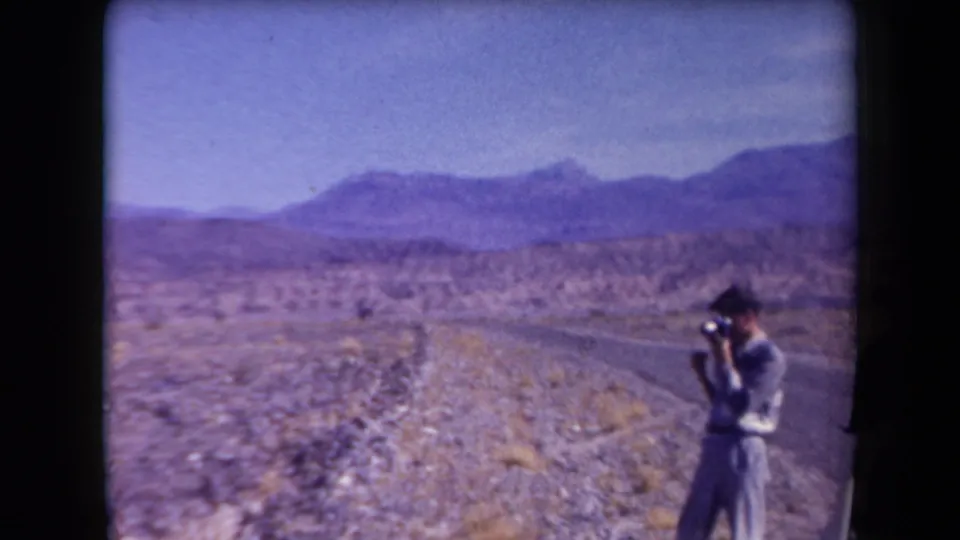 A man with a camera stands on a narrow Arizona desert road, framed by scrub, rocky ground, and distant mountains.