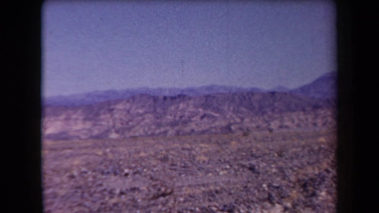 1958 Arizona 8mm frame showing rocky desert ground, distant ridges, and a soft blue-purple sky.