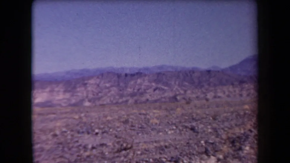 1958 Arizona 8mm frame showing rocky desert ground, distant ridges, and a soft blue-purple sky.