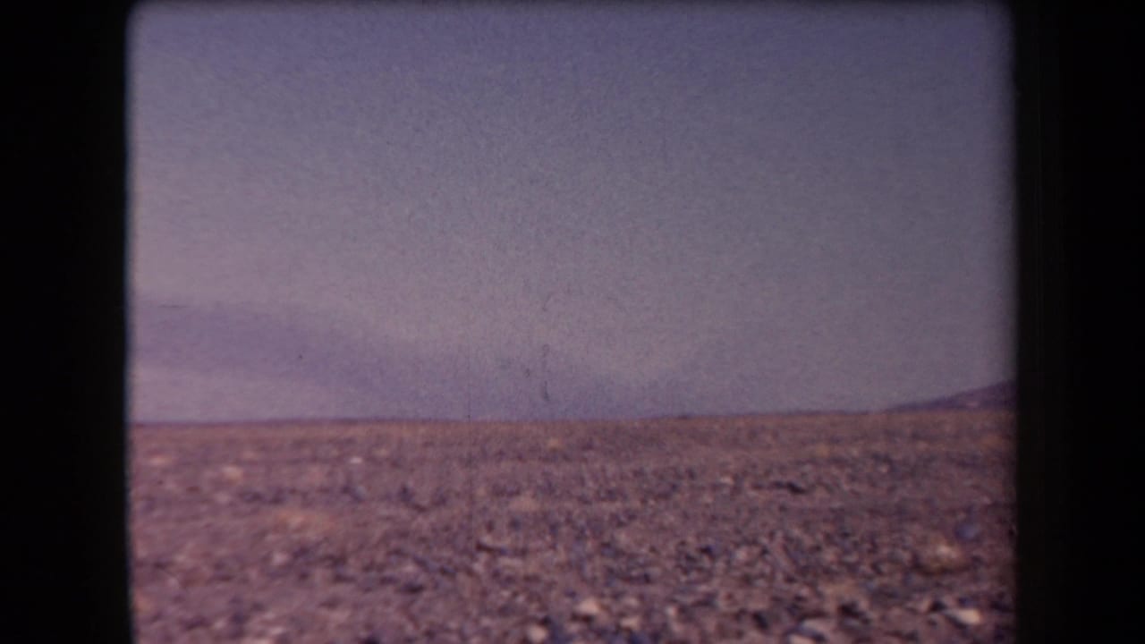1958 Arizona 8mm frame showing rocky desert ground, sparse brush, a hazy sky, and distant low mountains.