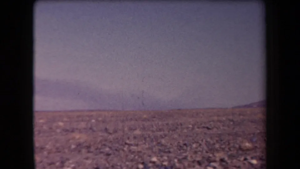 1958 Arizona 8mm frame showing rocky desert ground, sparse brush, a hazy sky, and distant low mountains.