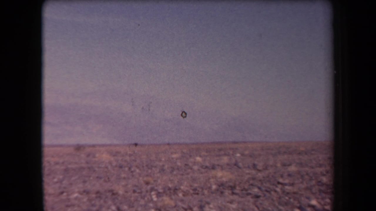 Open desert terrain with a hazy sky, distant mountains, and a small dark speck near the horizon.