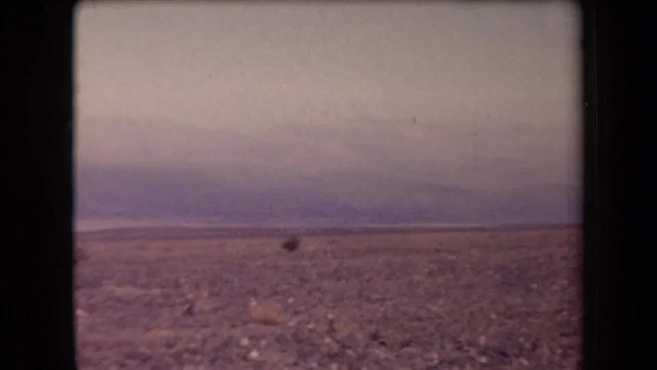 1958 Arizona 8mm frame with rocky desert terrain, hazy distant mountains, and a small dark distant object.