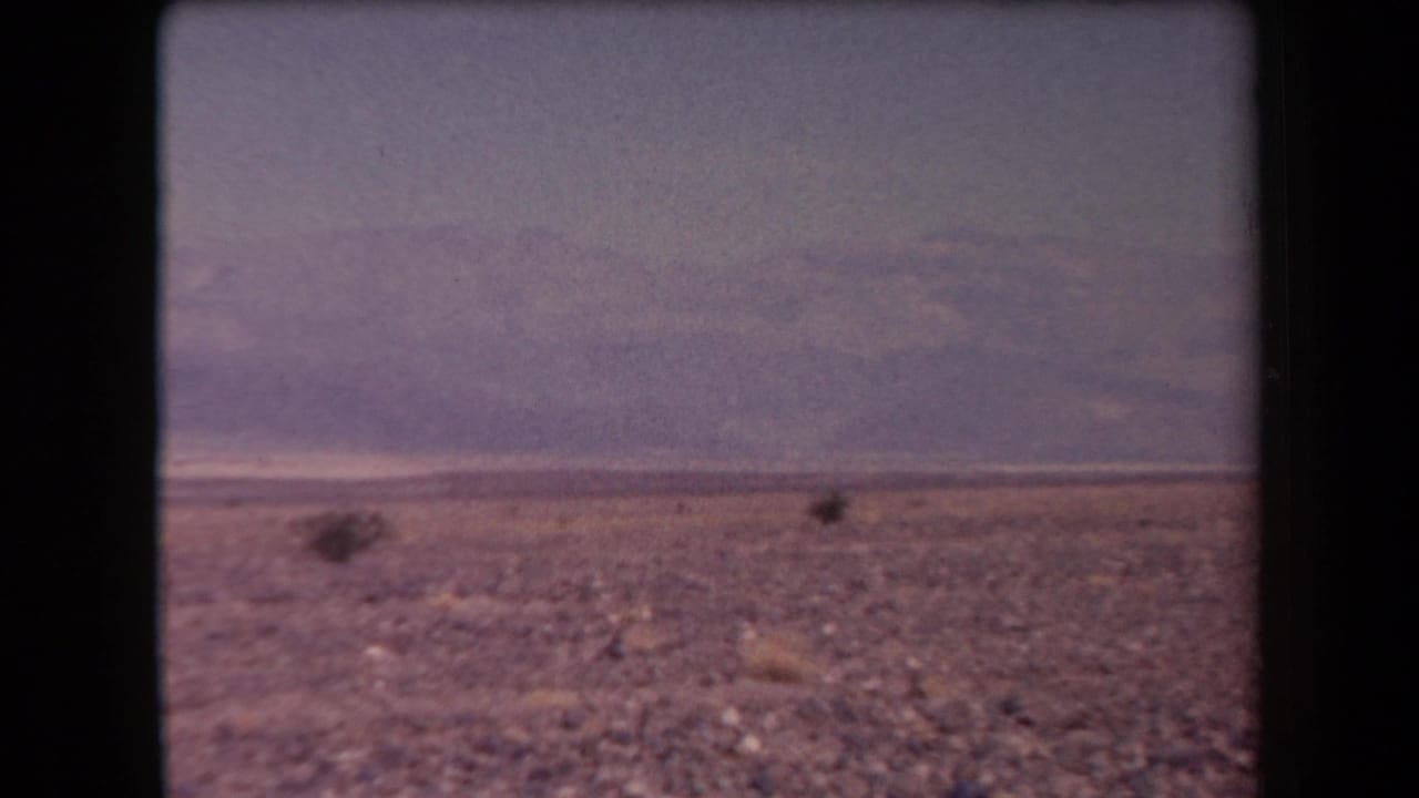 1958 Arizona 8mm frame showing gravelly desert ground, low shrubs, distant mountains, and a muted hazy sky.