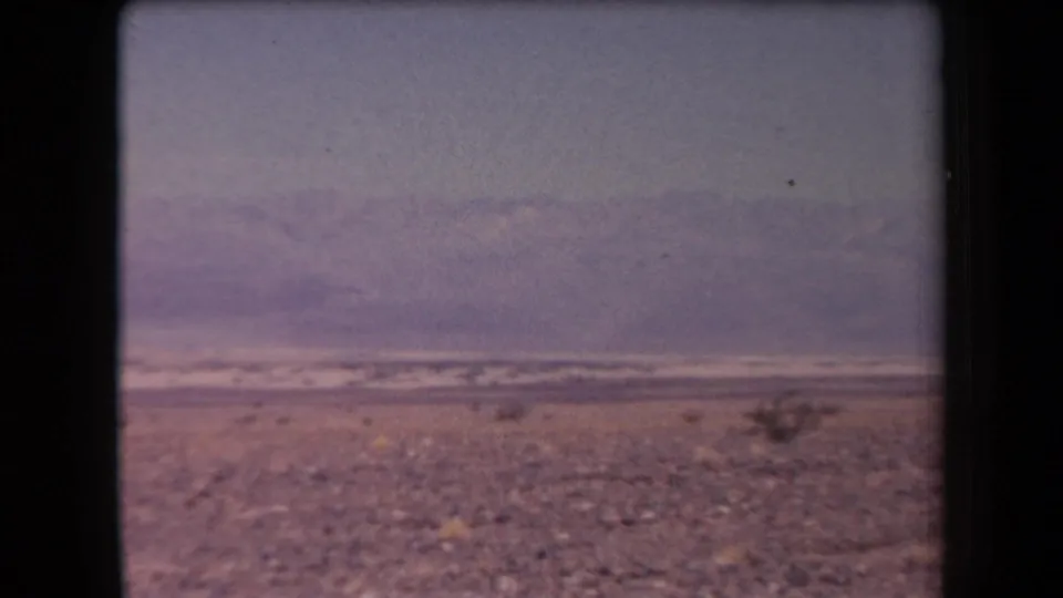 1958 Arizona 8mm frame showing sparse desert vegetation across a gravel plain with hazy mountains beyond.