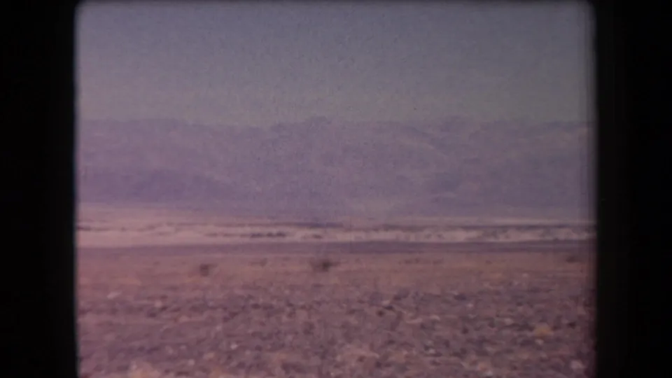 1958 Arizona 8mm frame of an open desert plain with sparse shrubs, rocky ground, distant mountains, and visible film grain.
