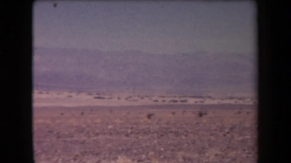 1958 Arizona 8mm frame showing a rocky desert plain, scattered scrub, and distant mountains through faded magenta haze.