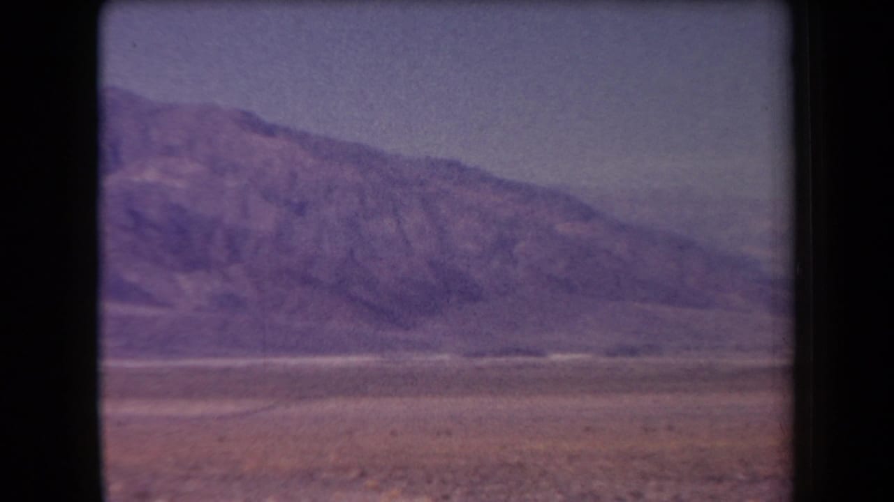 1958 Arizona 8mm frame showing a distant mountain range across an open desert plain with faded purple color and black reel borders.