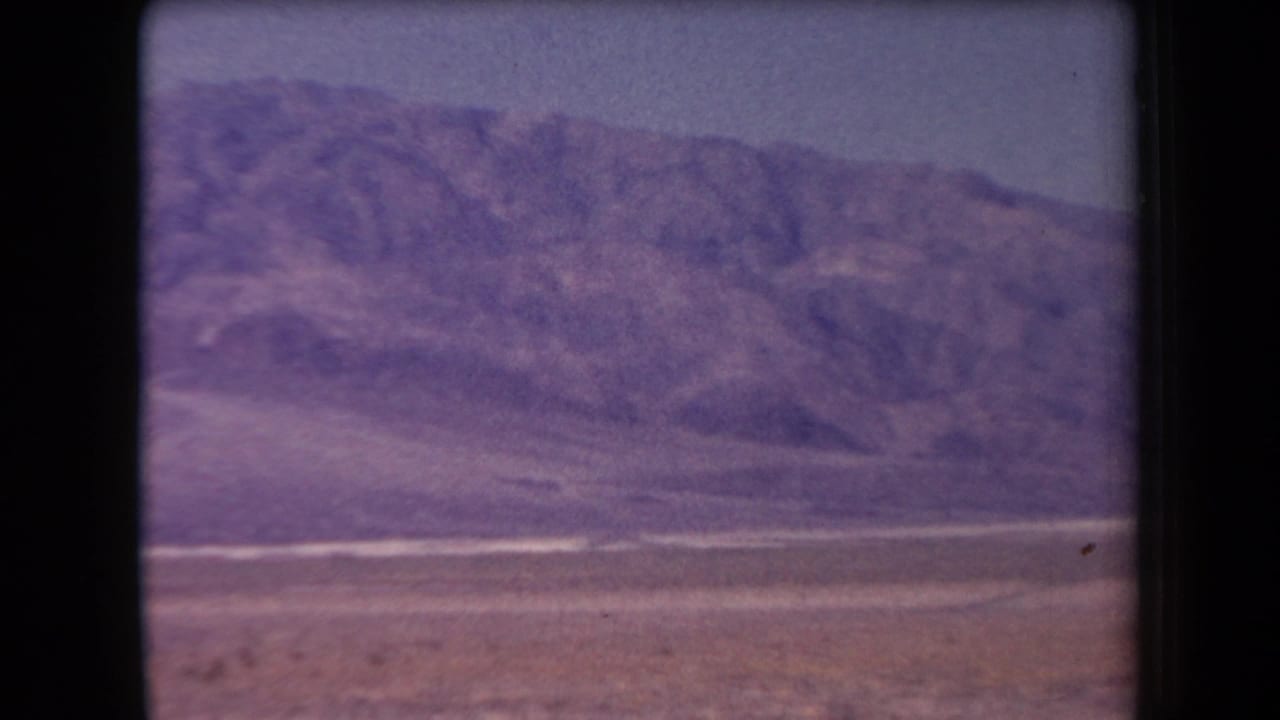 Rocky mountain slope seen across a dry valley floor with pale horizontal bands.