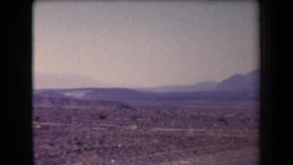 1958 Arizona 8mm frame showing rocky desert foreground, scattered scrub, and distant mountain ridges beneath open sky.