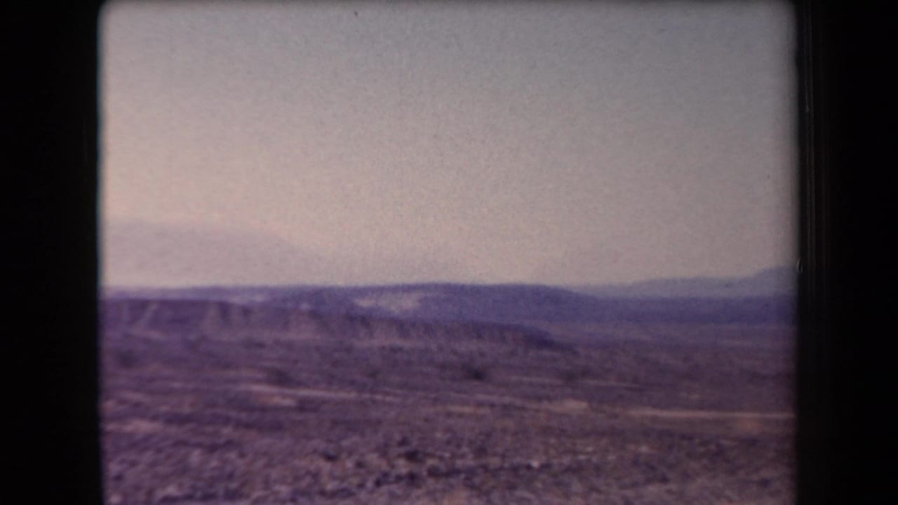 1958 Arizona 8mm frame showing scrubby desert ground, low ridges, and distant mountains beneath a hazy sky.