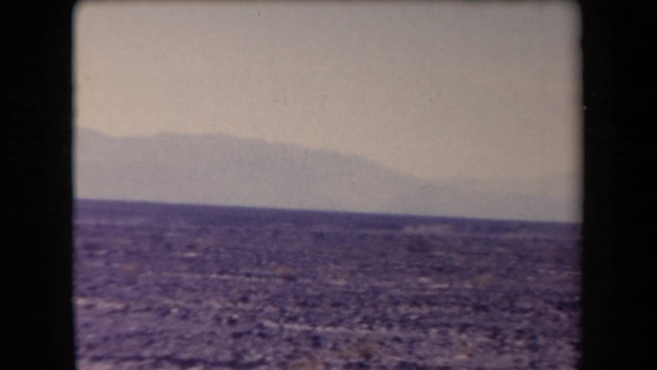 1958 Arizona 8mm frame showing open desert ground, low horizon, distant hazy mountains, and a pale sky.