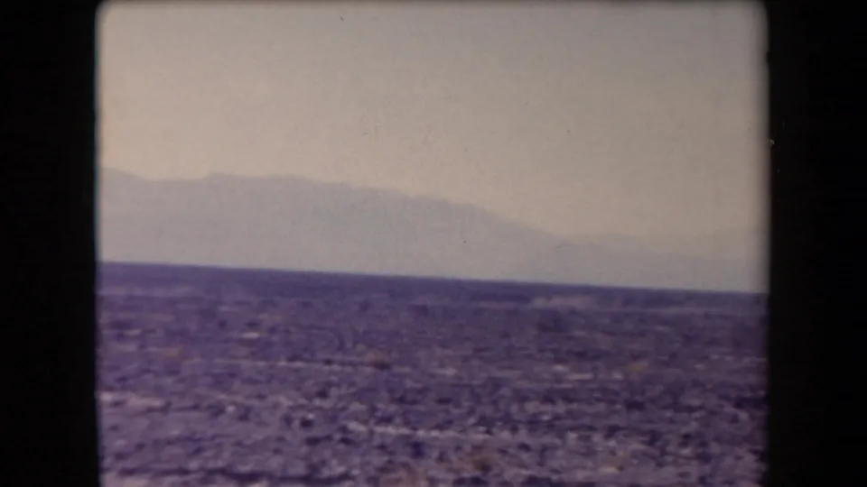 1958 Arizona 8mm frame showing open desert ground, low horizon, distant hazy mountains, and a pale sky.