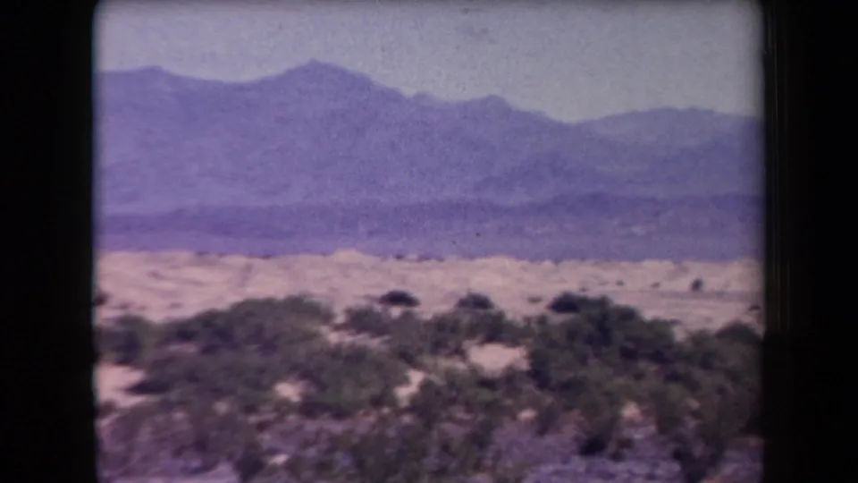 Vintage Arizona 8mm frame with low shrubs, pale desert floor, and a hazy mountain range under a washed sky.