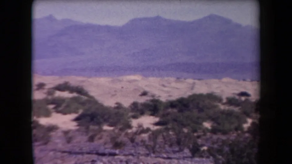 1958 Arizona home-movie frame showing rocky foreground, scattered desert shrubs, and distant mountains through color-shifted film.