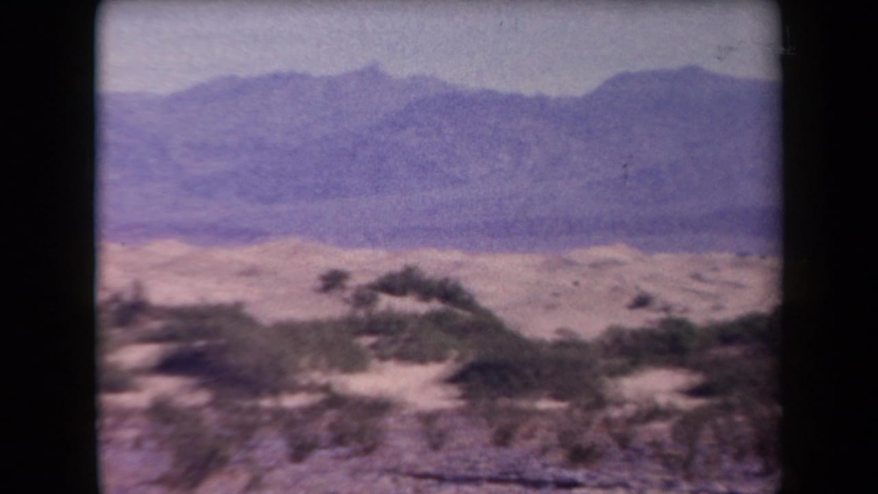 1958 Arizona 8mm frame showing low desert shrubs, sandy ground, and distant mountain ridges with faded purple color.