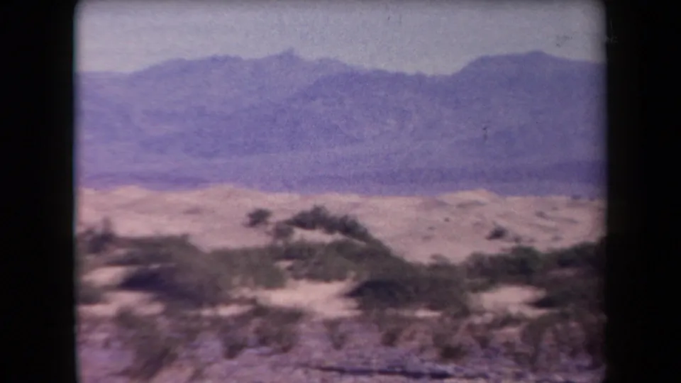 1958 Arizona 8mm frame showing low desert shrubs, sandy ground, and distant mountain ridges with faded purple color.