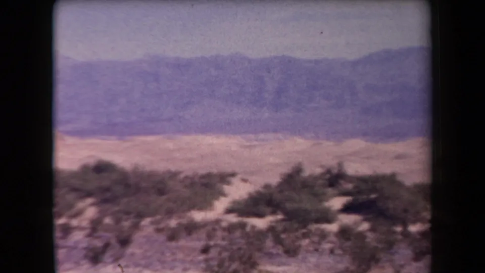 Arizona desert landscape in a faded 1958 8mm home-movie frame, with scrub plants, pale sand, and a distant ridge.