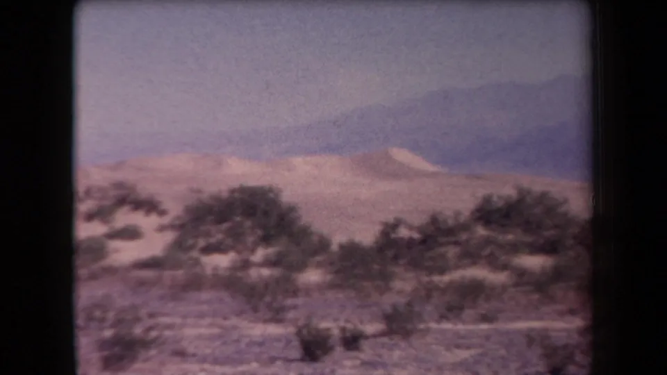 Faded 1958 Arizona 8mm frame of scrub vegetation, sandy desert ground, and a broad hazy mountain slope.