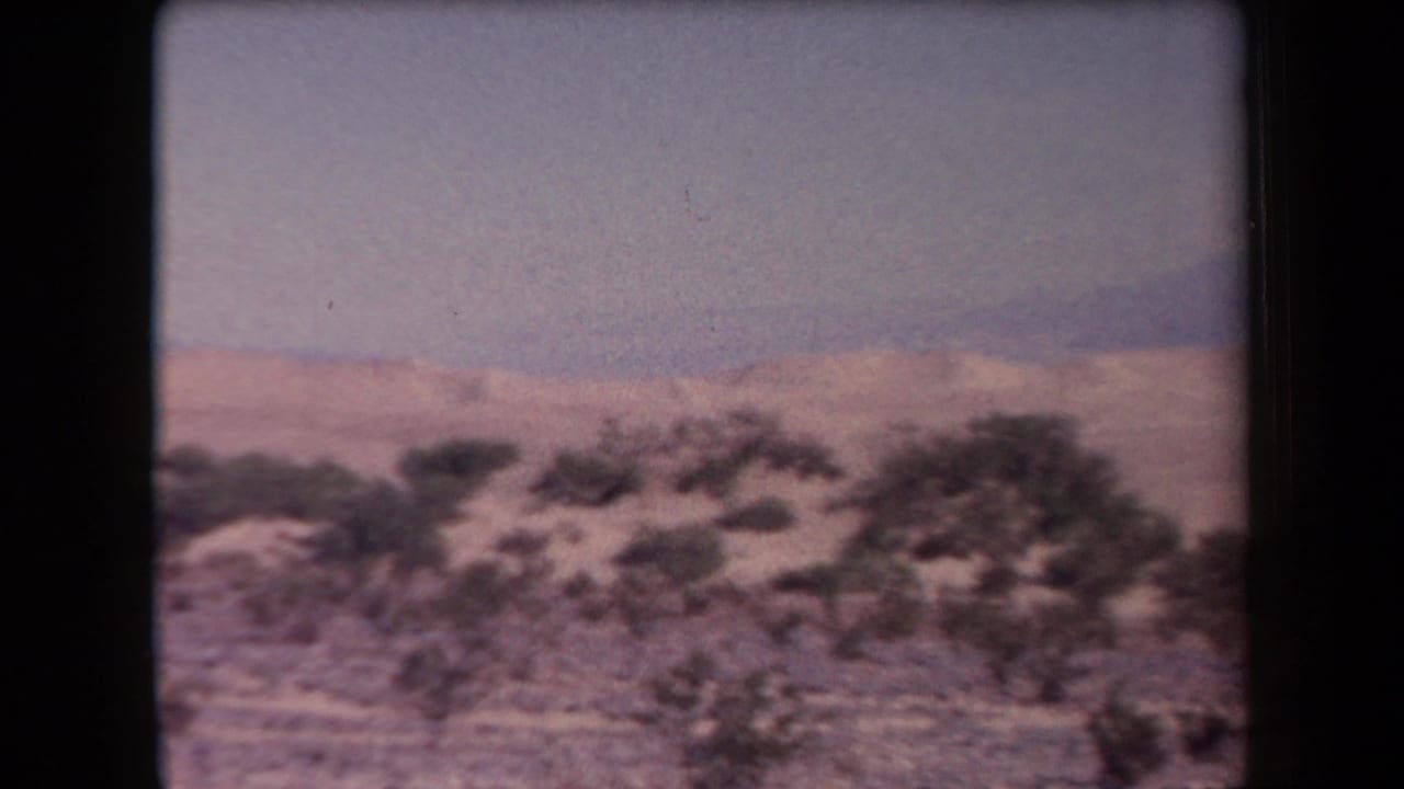 1958 Arizona home-movie frame showing scrubby desert terrain, pale sand, distant mountains, and a hazy sky during a handheld pan.