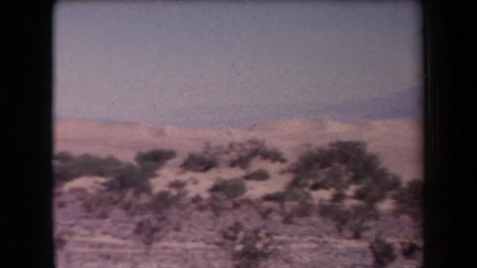 1958 Arizona home-movie frame showing scrubby desert terrain, pale sand, distant mountains, and a hazy sky during a handheld pan.