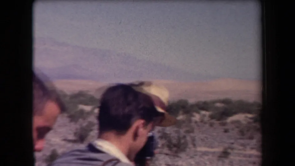 1958 Arizona roadside view with adult travelers in casual clothing and a hat partly framed against desert scrub and distant mountains.