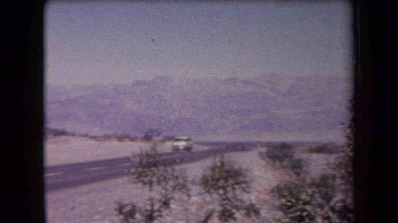 1958 Arizona home-movie frame showing a paved desert highway, sparse roadside vegetation, distant mountains, and a vehicle on the road.