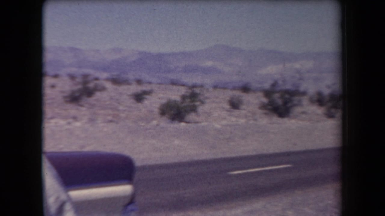 1958 Arizona roadside view with a partial two-tone sedan at the edge of frame, a paved highway, desert scrub, and distant mountains.