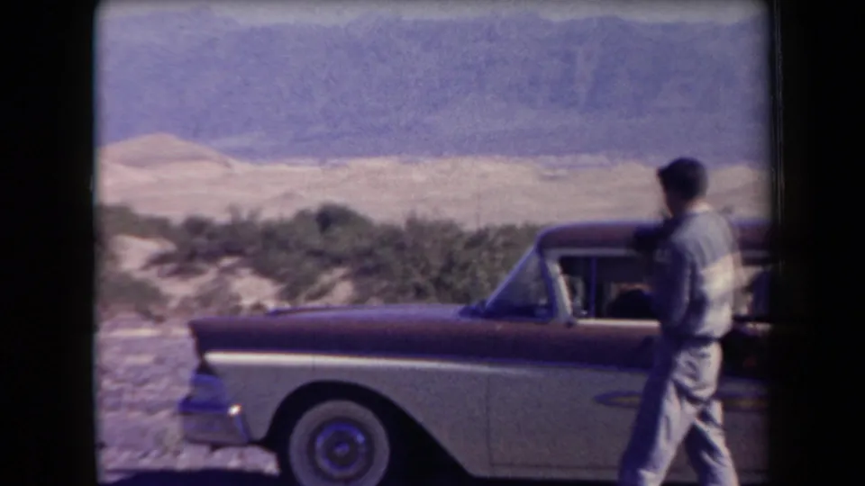 1958 Arizona frame showing an adult man near a parked two-tone 1950s sedan with desert scrub and pale mountains in the background.