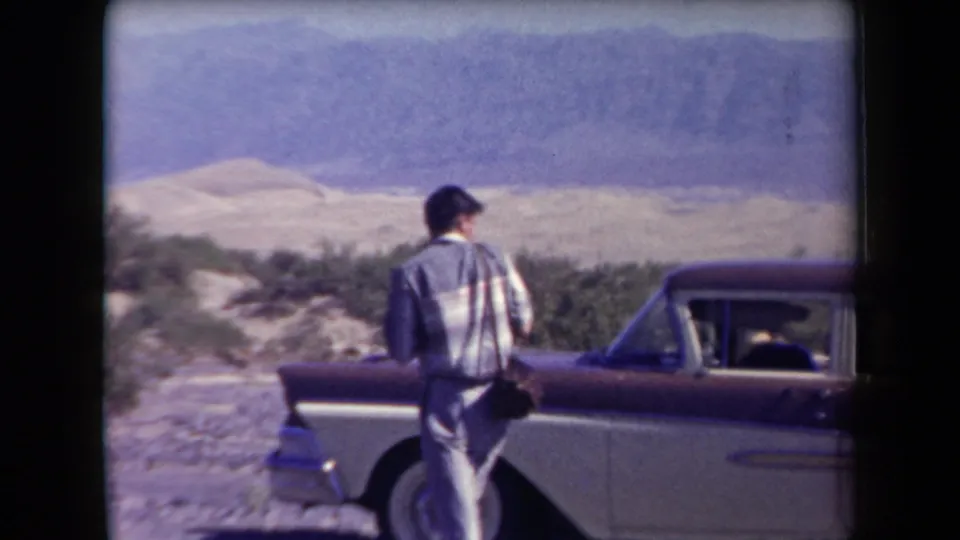 1958 Arizona home-movie frame with an adult man seen from behind near a two-tone sedan, desert vegetation, and mountains beyond.