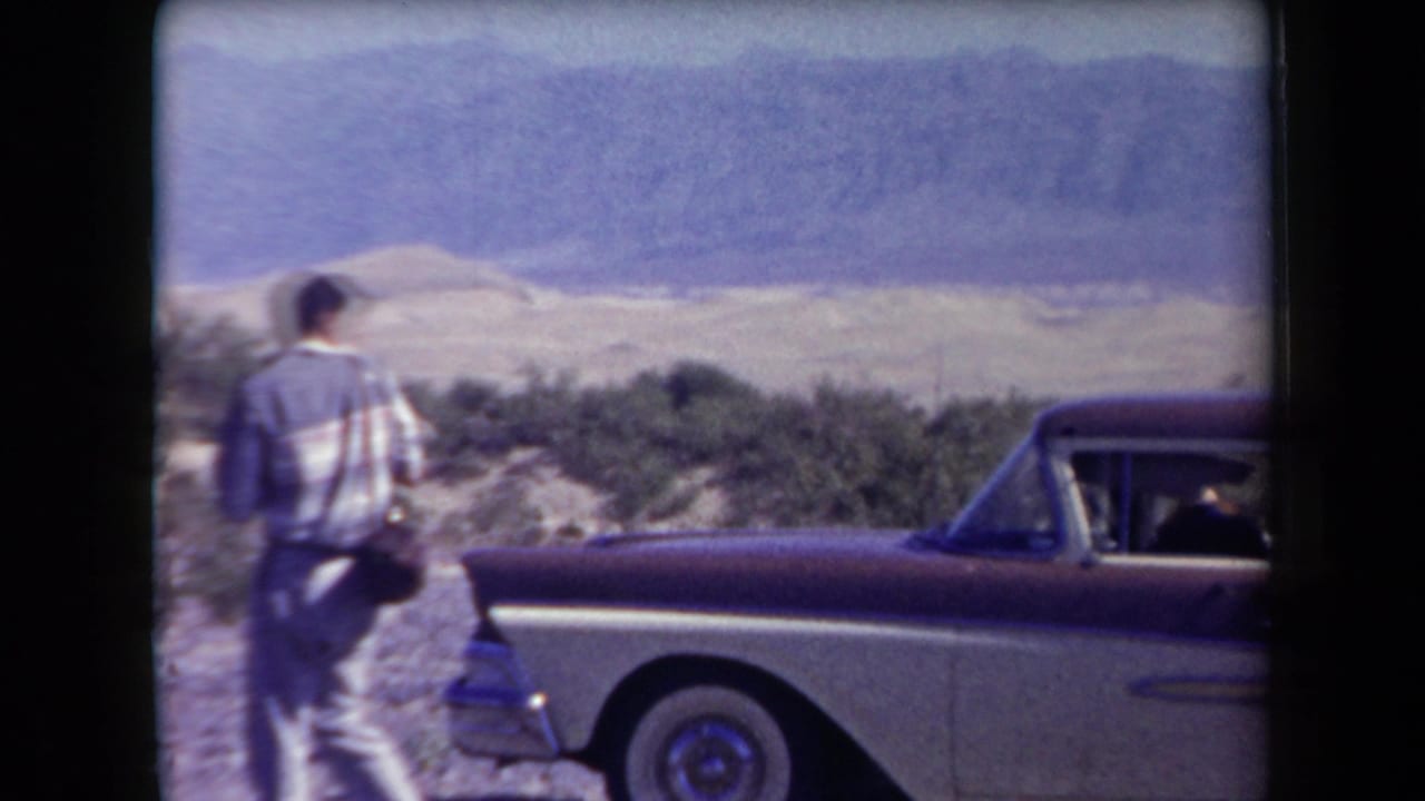 1958 Arizona home-movie frame showing a man beside a two-tone car with scrub, sand dunes, and mountains beyond.