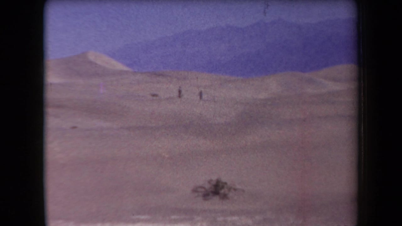 Small figures walk across pale desert dunes with mountains in the background.