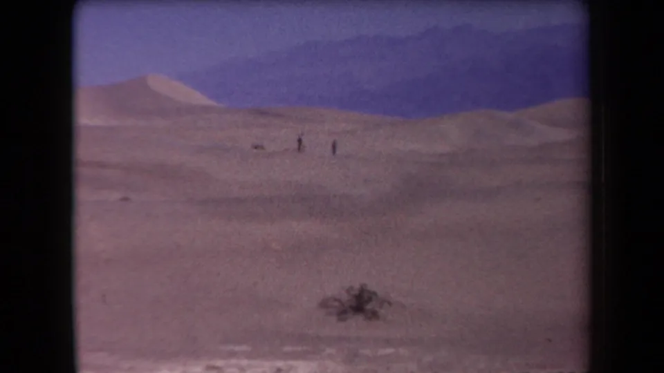 Wide 1958 Arizona home-movie view of dunes, distant people, foreground scrub, and mountain silhouettes.