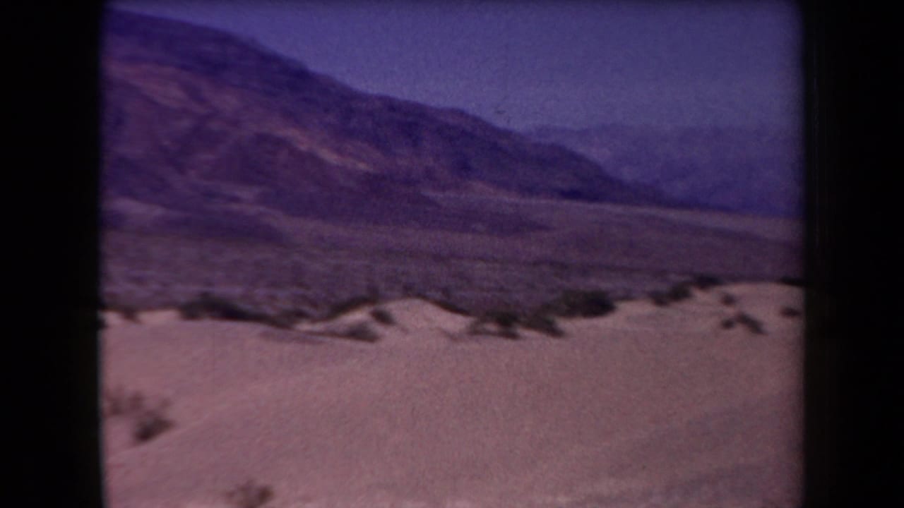 1958 Arizona 8mm frame showing pale desert ground, scattered scrub, a wide valley floor, and distant rugged mountains with aged purple color shift.