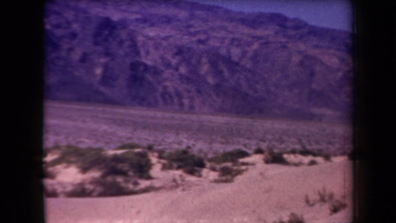 1958 Arizona 8mm view of sparse desert vegetation, a flat valley basin, and rugged mountain slopes, framed by dark film edges.