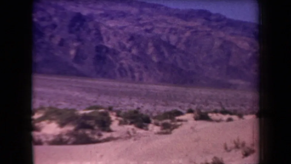 1958 Arizona 8mm view of sparse desert vegetation, a flat valley basin, and rugged mountain slopes, framed by dark film edges.