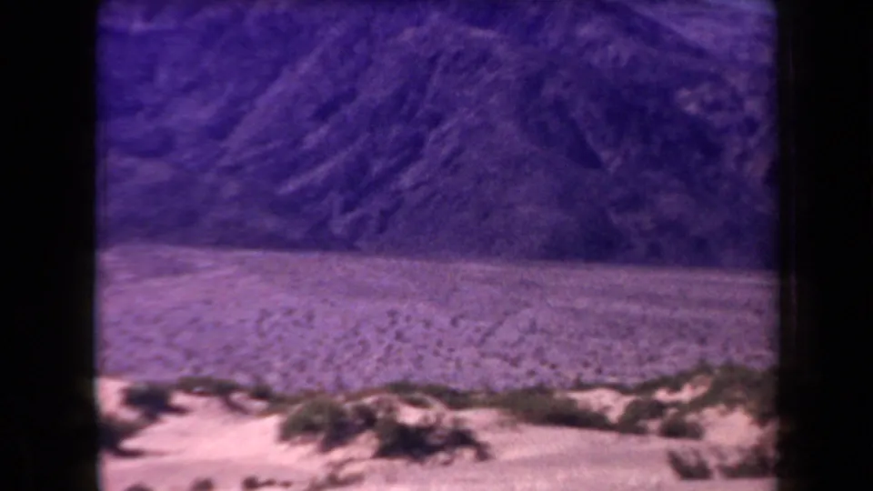1958 Arizona home-movie frame with foreground scrub, patterned desert floor, and steep mountain ridges in faded purple-blue color.