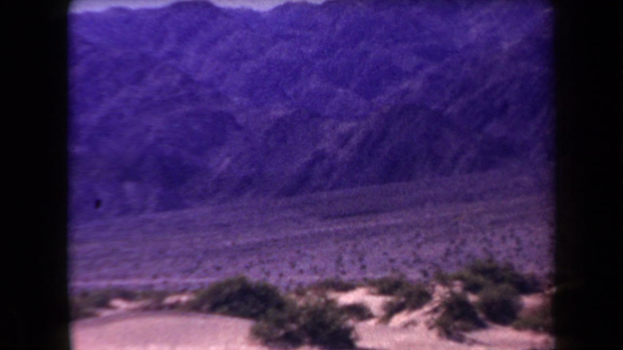 1958 Arizona 8mm frame showing sandy desert brush in front of distant mountains, with soft focus and purple color shift.