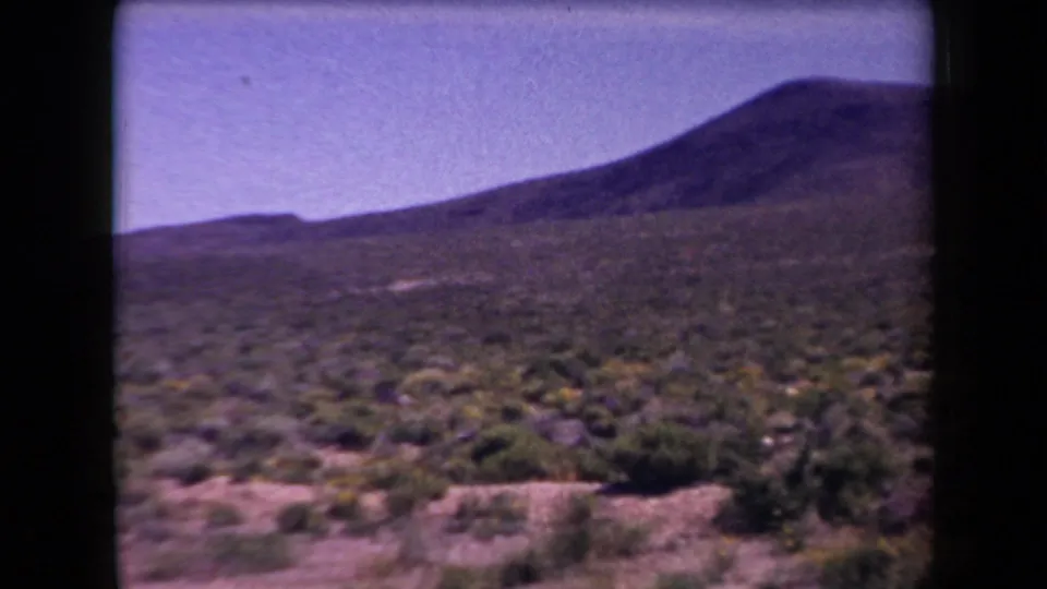 1958 Arizona 8mm frame showing low desert brush across a flat plain, with a dark rounded hill in the distance.