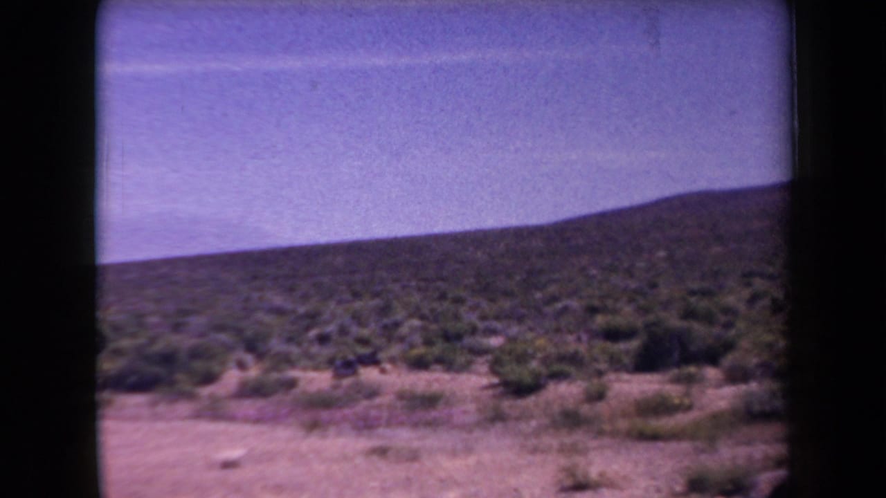 1958 Arizona 8mm frame showing open scrub desert and a low hillside under a pale blue sky near a highway shoulder.