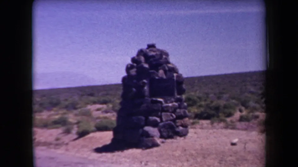 1958 Arizona roadside view with a small stacked-stone monument set against scrub brush and open desert sky.
