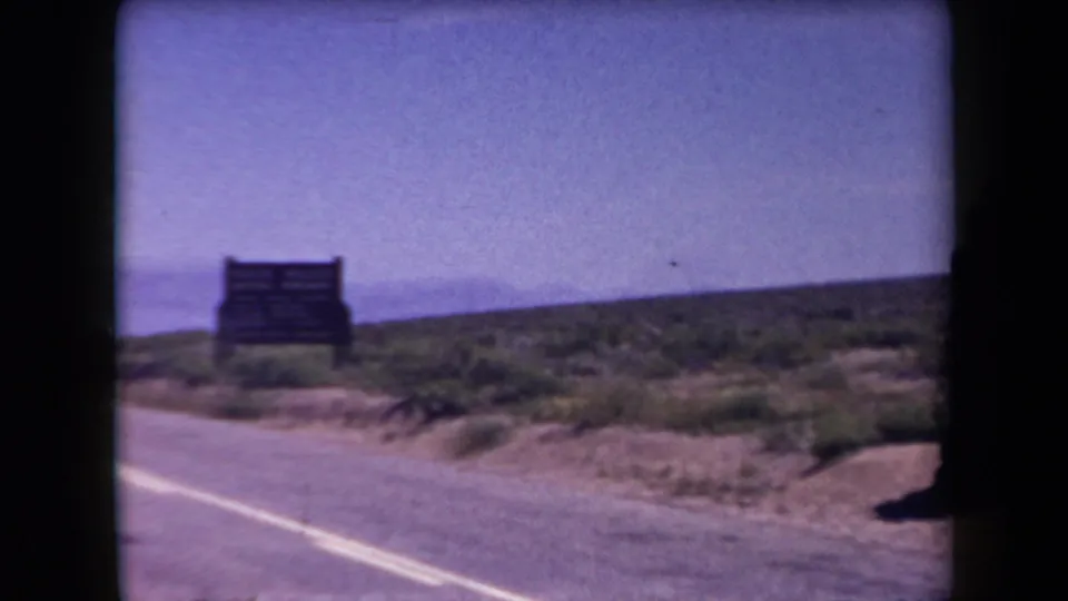 1958 Arizona frame showing a wooden roadside sign beside a paved highway and scrub-covered desert terrain.