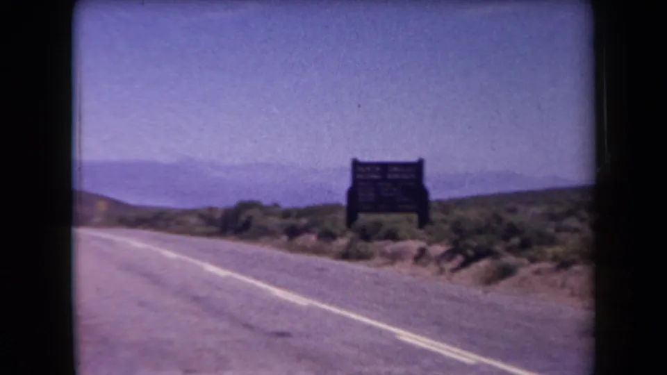 1958 Arizona 8mm frame of a quiet paved highway, wooden roadside sign, scrub desert, and hazy distant mountains.