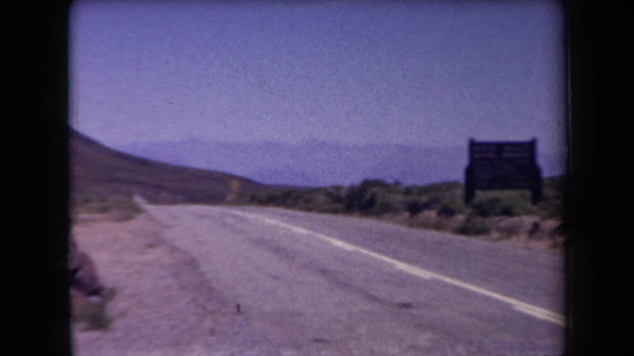 1958 Arizona home-movie frame showing a paved desert road, arid roadside brush, a dark sign, and distant mountains under a hazy sky.