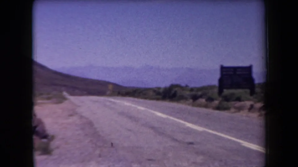 1958 Arizona home-movie frame showing a paved desert road, arid roadside brush, a dark sign, and distant mountains under a hazy sky.