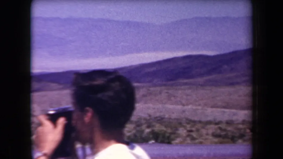 A man in the 1958 Arizona travel footage raises a small camera toward a wide desert and mountain landscape.