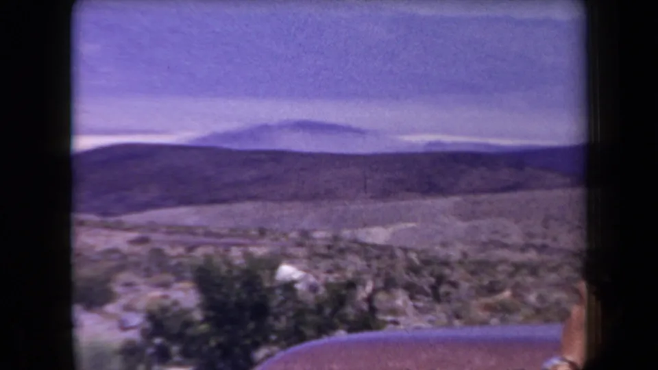 1958 Arizona 8mm frame of a broad desert valley, low mountains, scrub vegetation, and a roadside guardrail under a hazy sky.