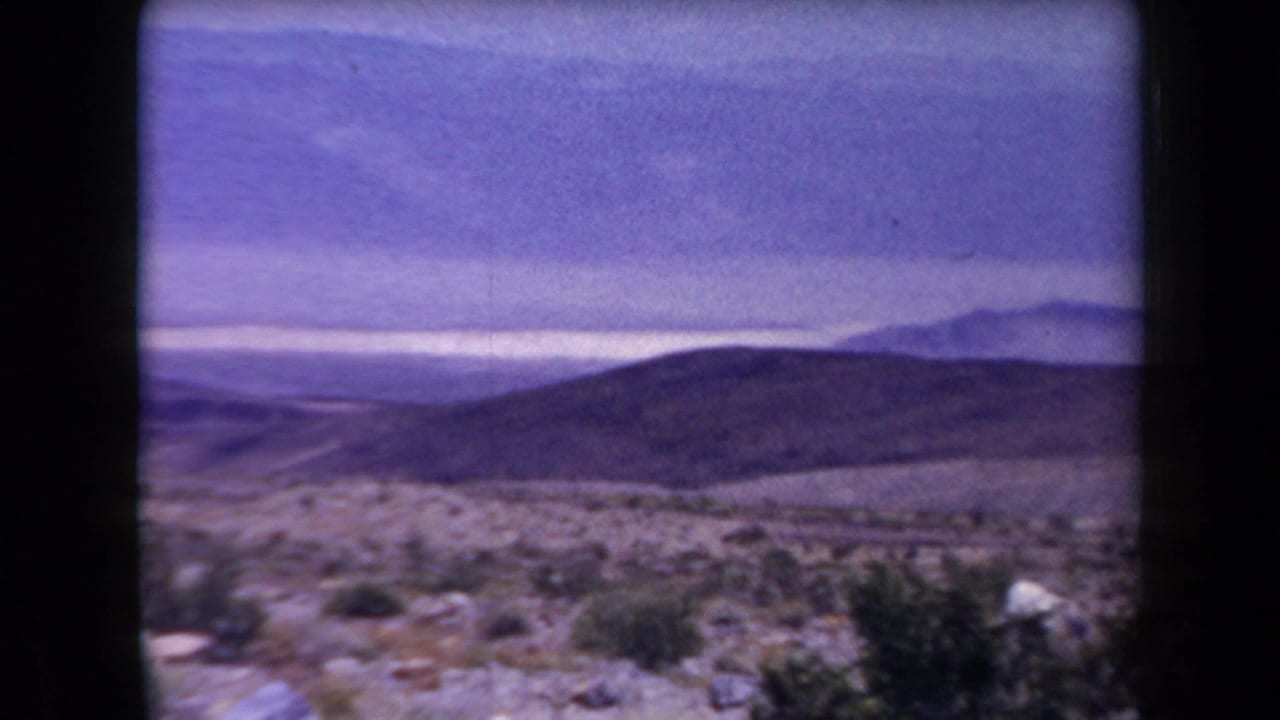 1958 Arizona 8mm frame showing rocky desert scrub, rolling hills, distant mountain ranges, and a hazy purple-tinted sky.